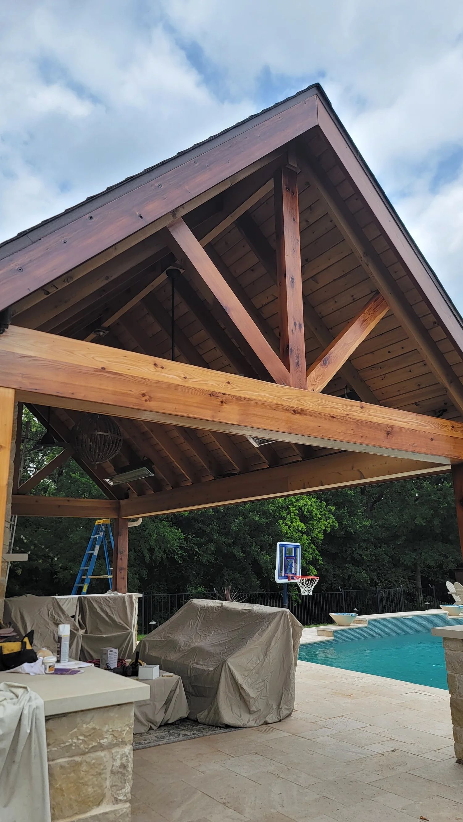 Wooden patio cover with a pool in the background. Construction in progress, cloudy sky.