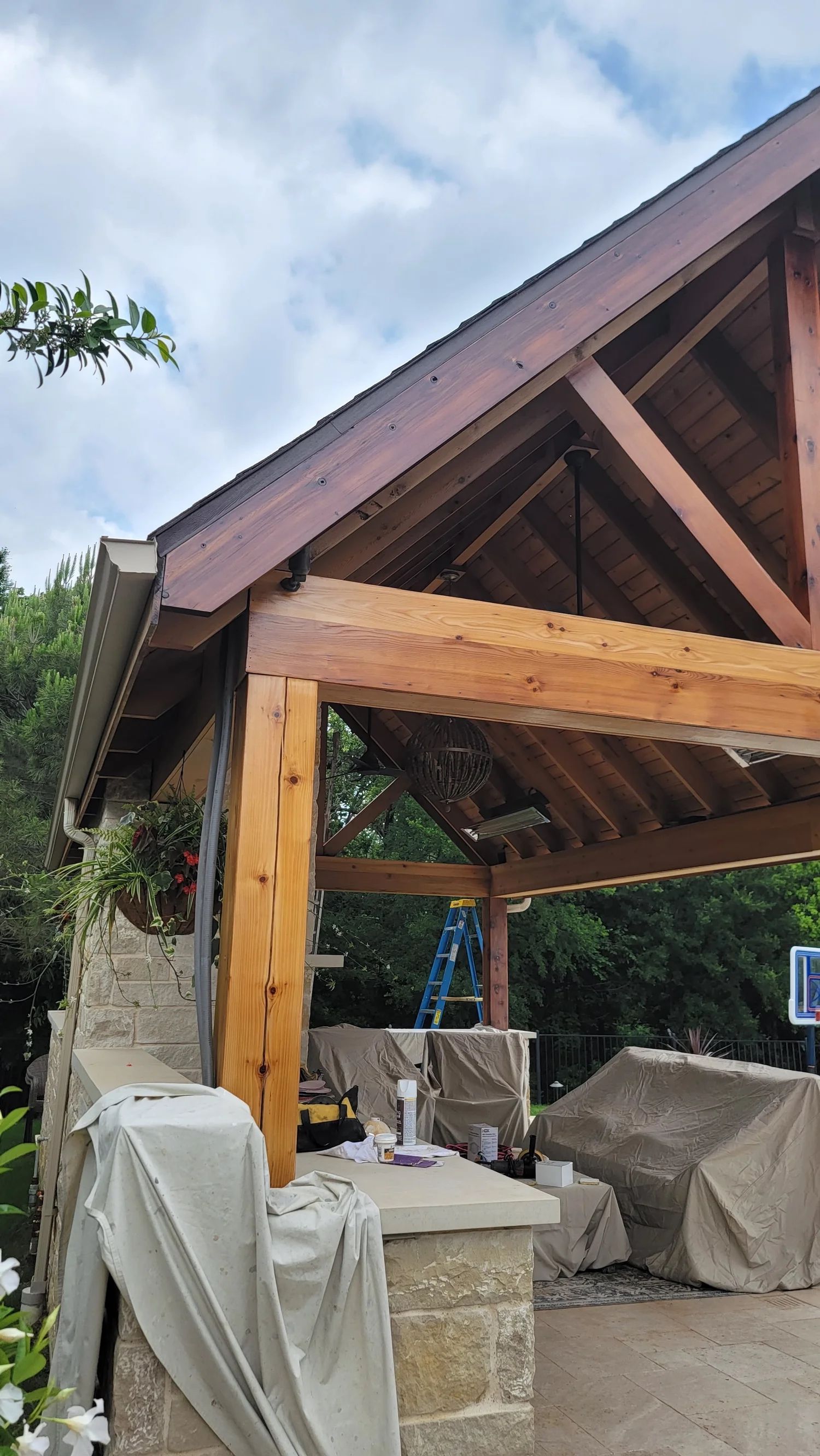 Wooden pavilion under construction, natural wood beams, stone wall, blue ladder, cloudy sky.
