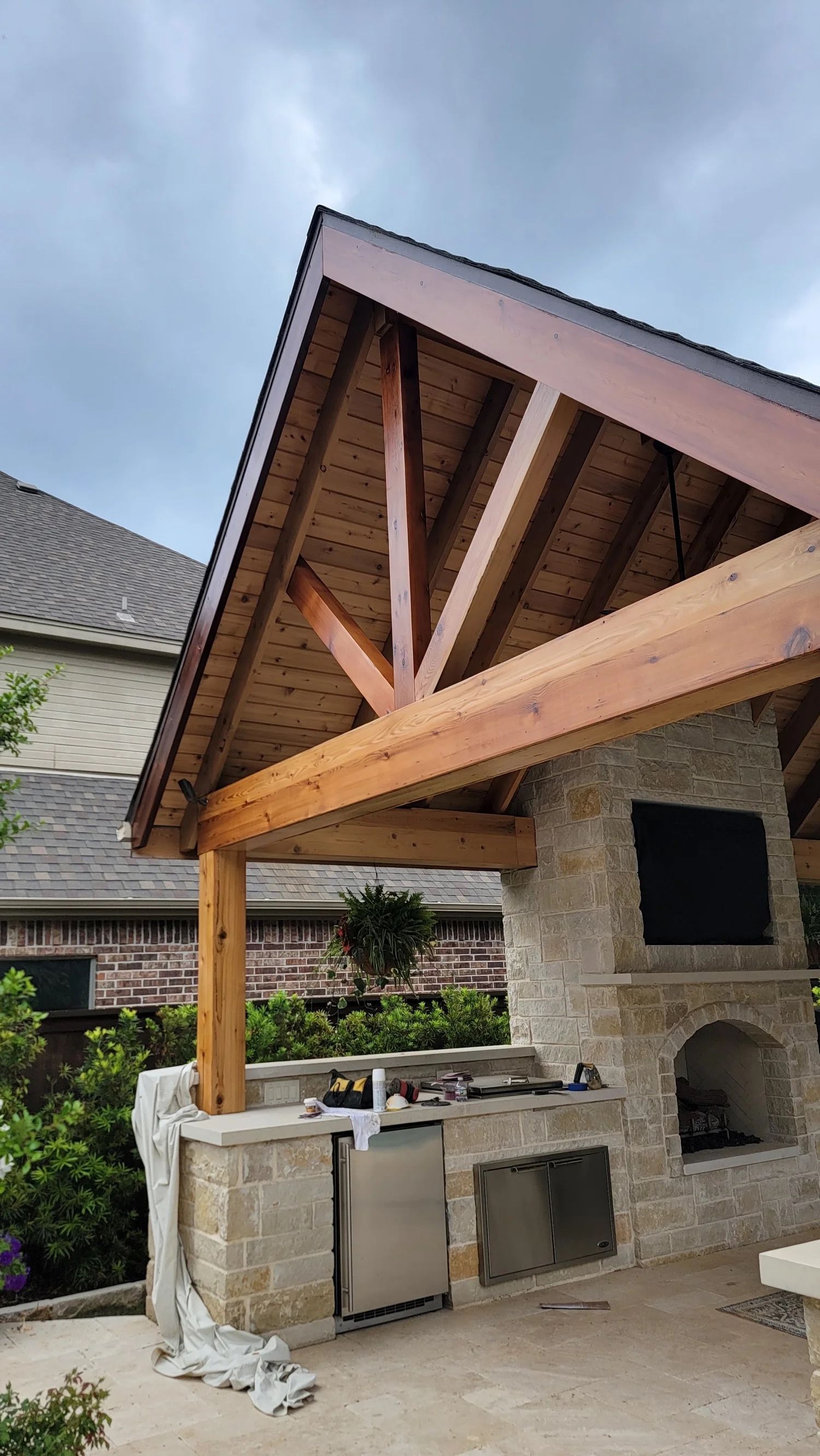 Outdoor kitchen with stone, wood, and a gabled roof. Includes a grill, sink, and fireplace.