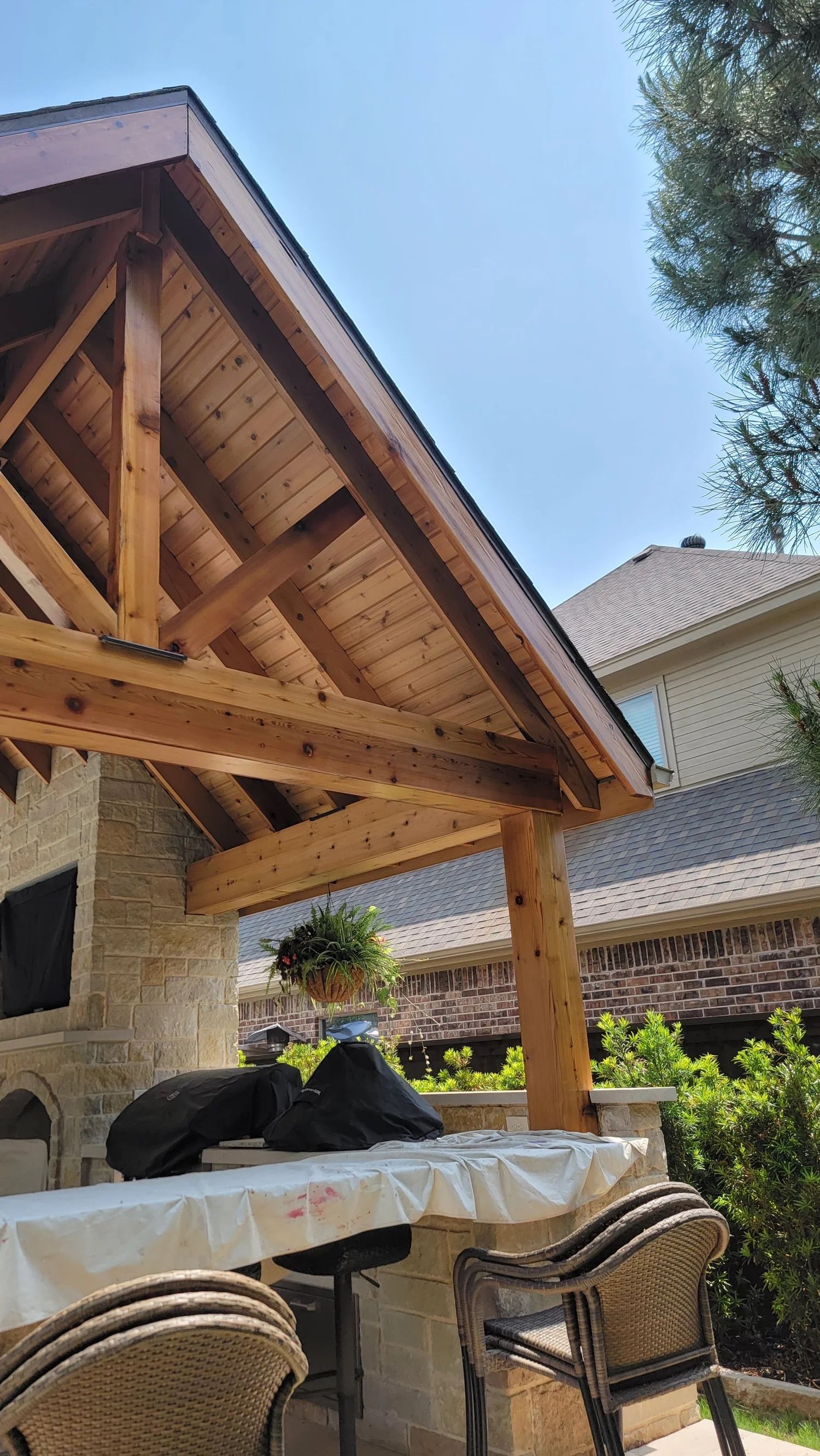 Wooden pergola over a stone outdoor kitchen with a bar, chairs, and a house in the background under a blue sky.