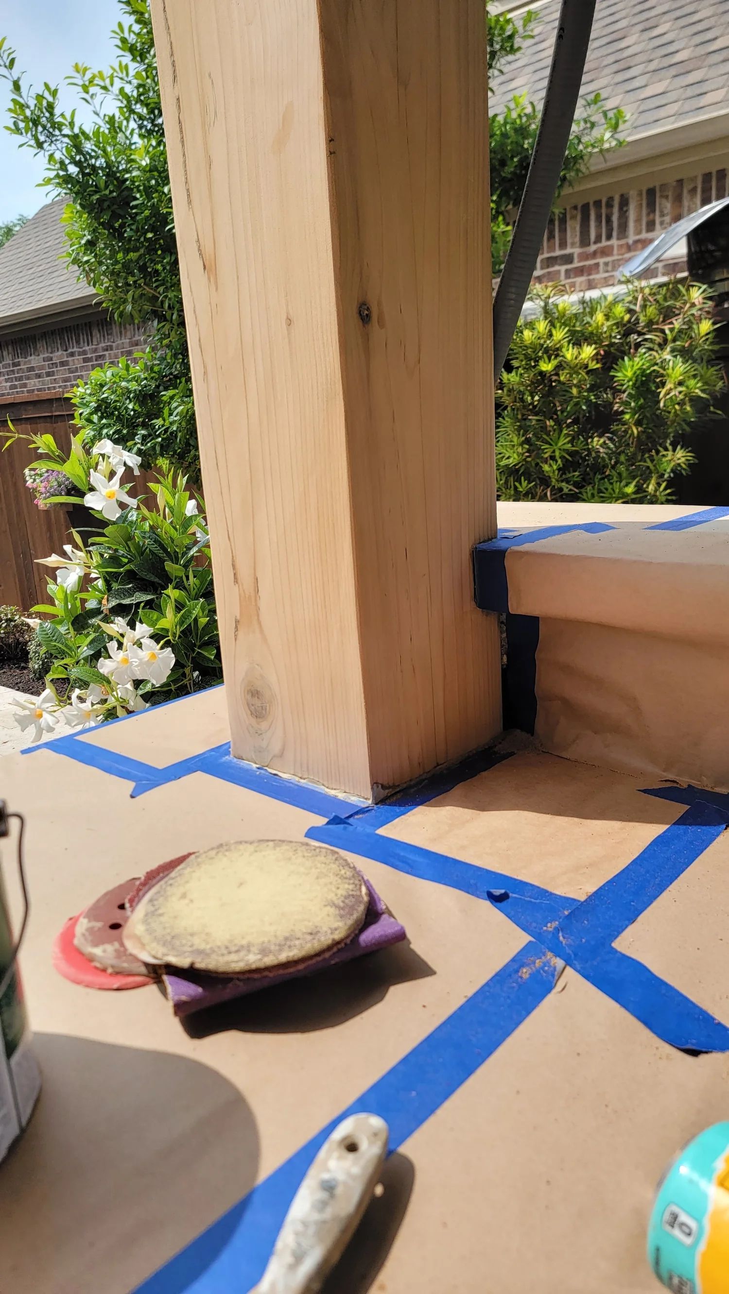Wooden post with painter's tape, sanding discs, and paint cans, preparing for staining on a patio.