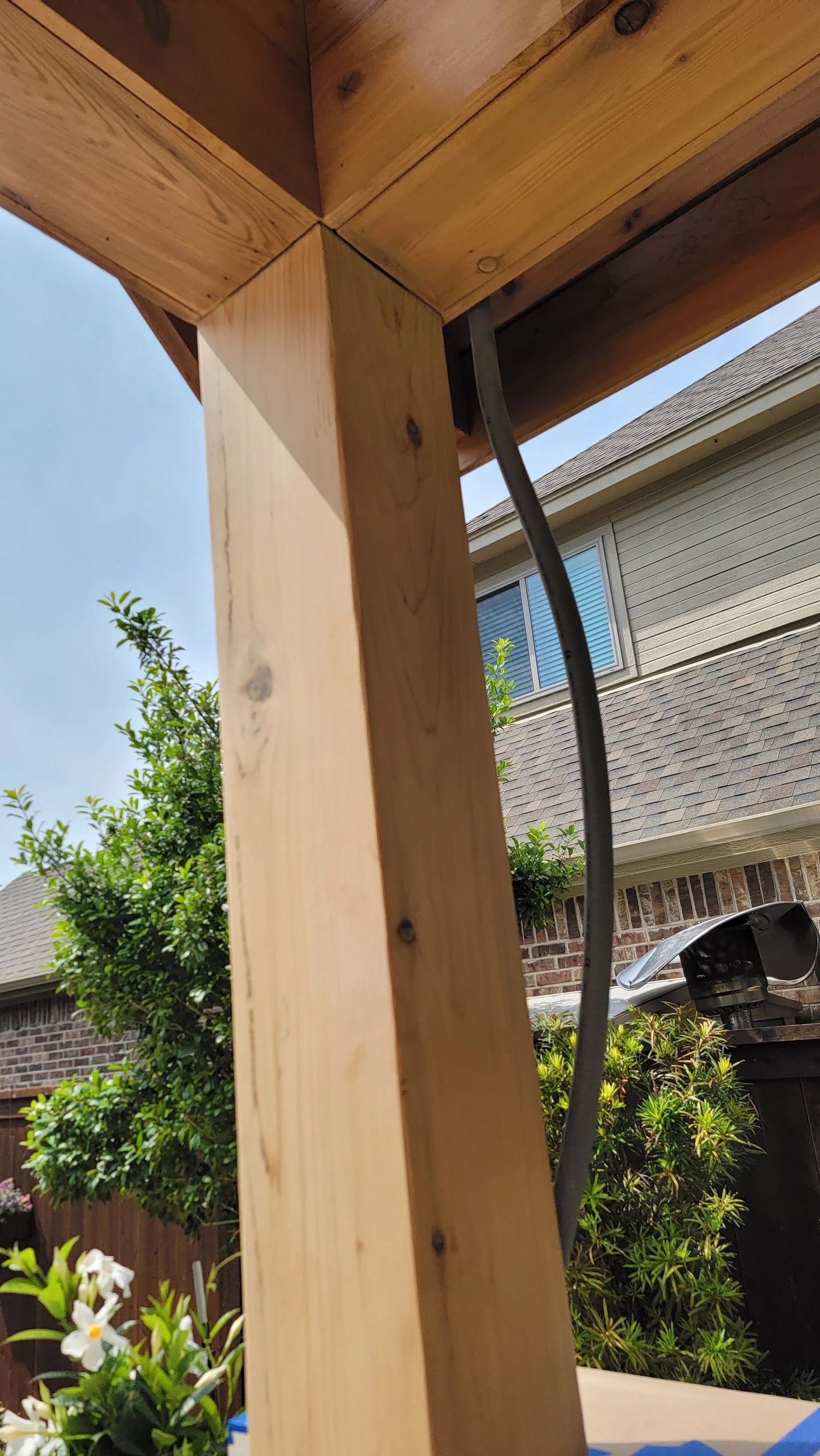 Wooden beam supports a corner of a structure. A grey cable runs along the beam's side; trees and houses are visible in the background.