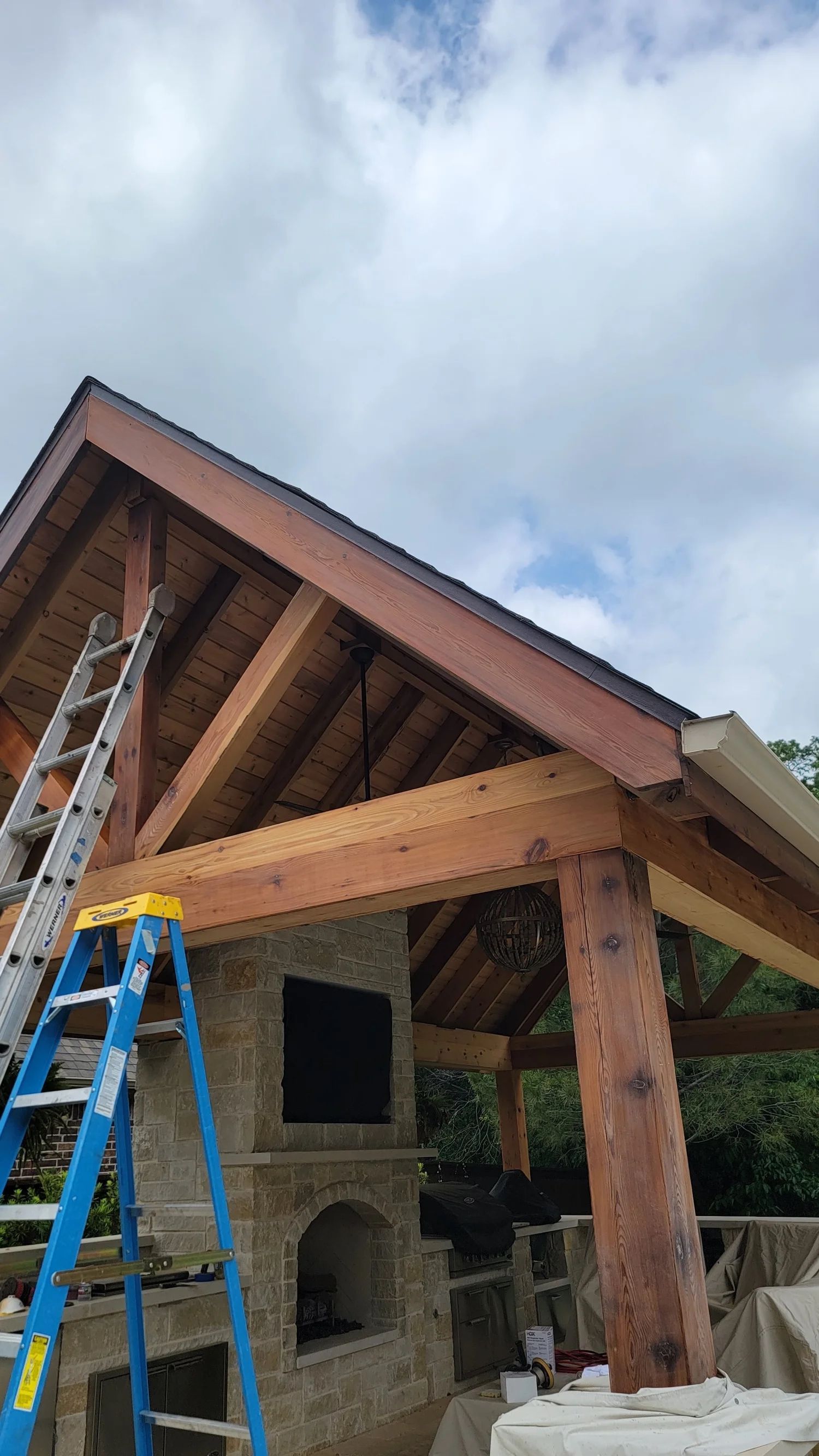 Outdoor wooden pavilion under construction with fireplace, blue ladder, and cloudy sky.