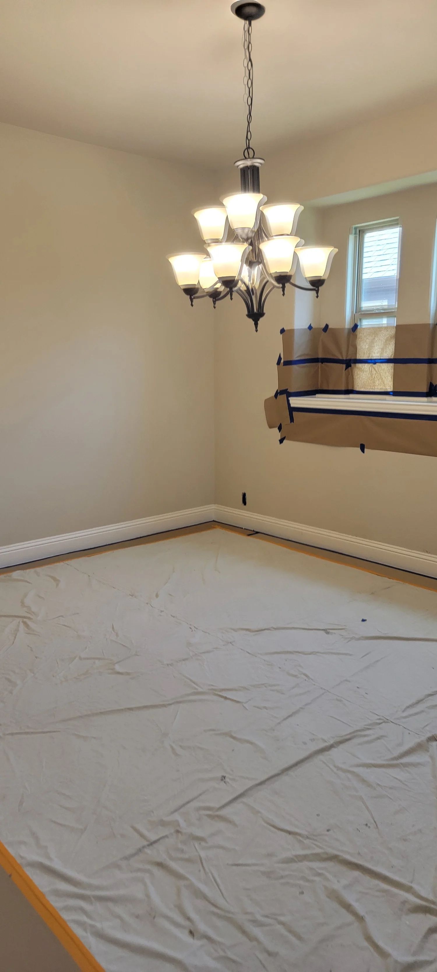 A chandelier hangs in a room with light-colored walls and a floor covered with a protective sheet.