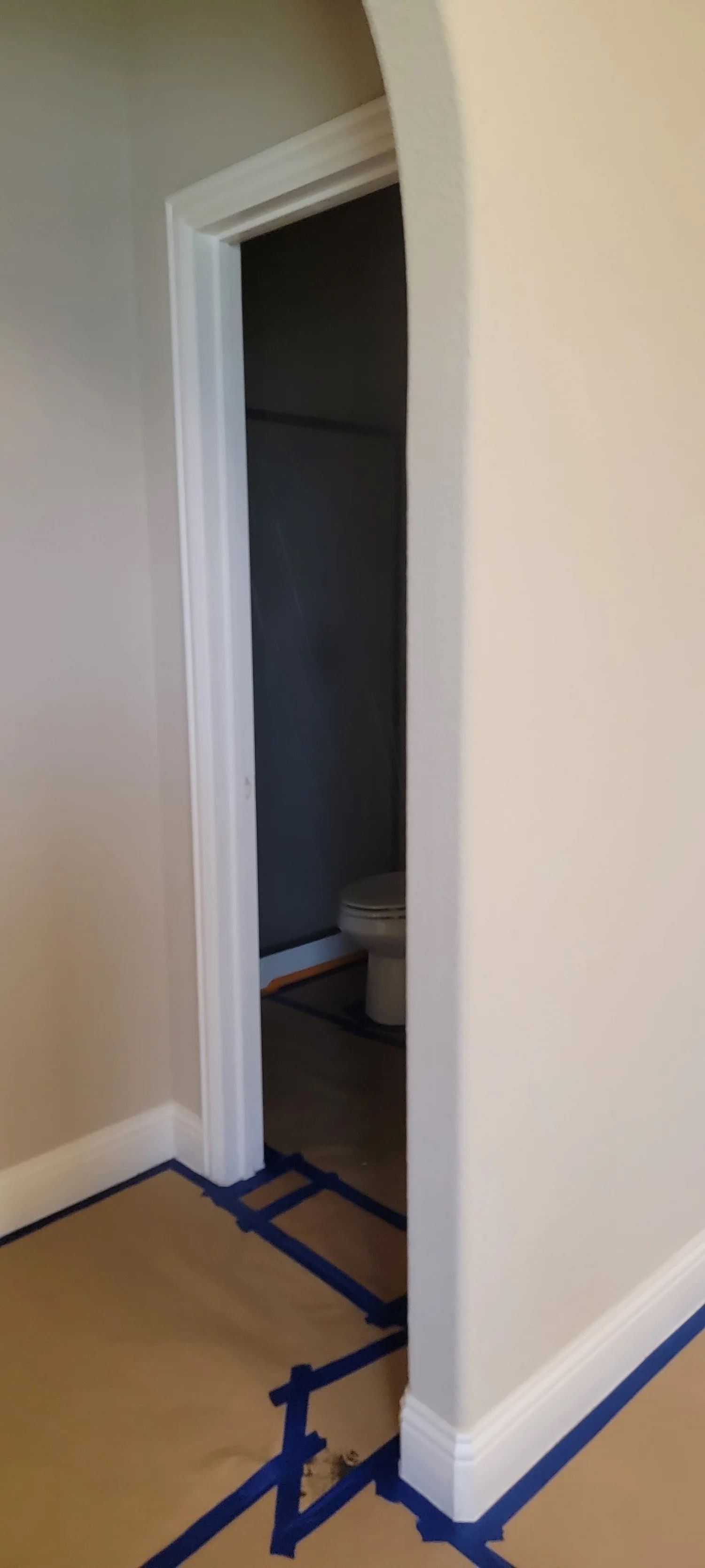 An archway leads into a room with a toilet. Walls are painted off-white; blue tape protects the floor during painting.