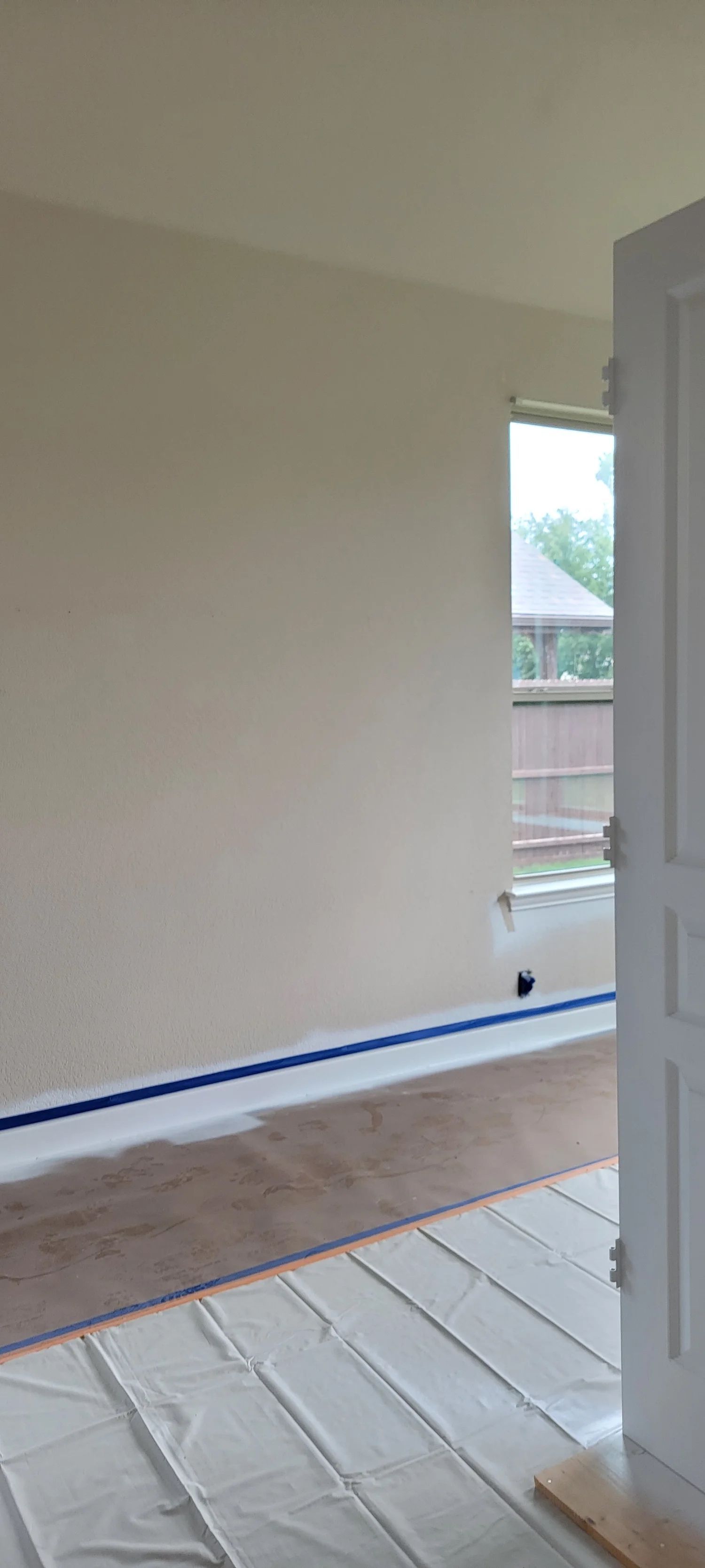 Interior room with beige walls, blue painter's tape, and protective floor covering. A door is open.