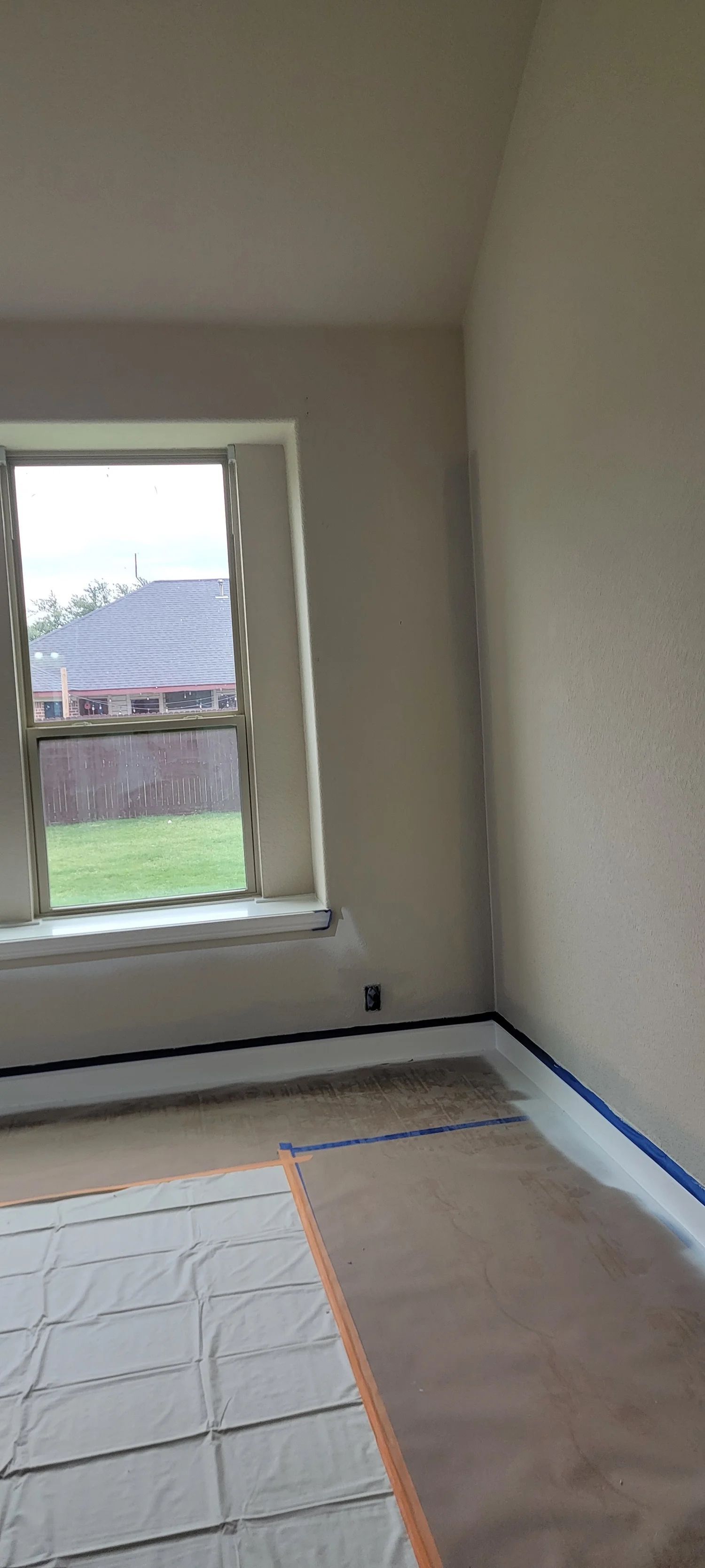 Interior of a room with a window, painting in progress. Walls are taped, floor covered, and house outside the window.