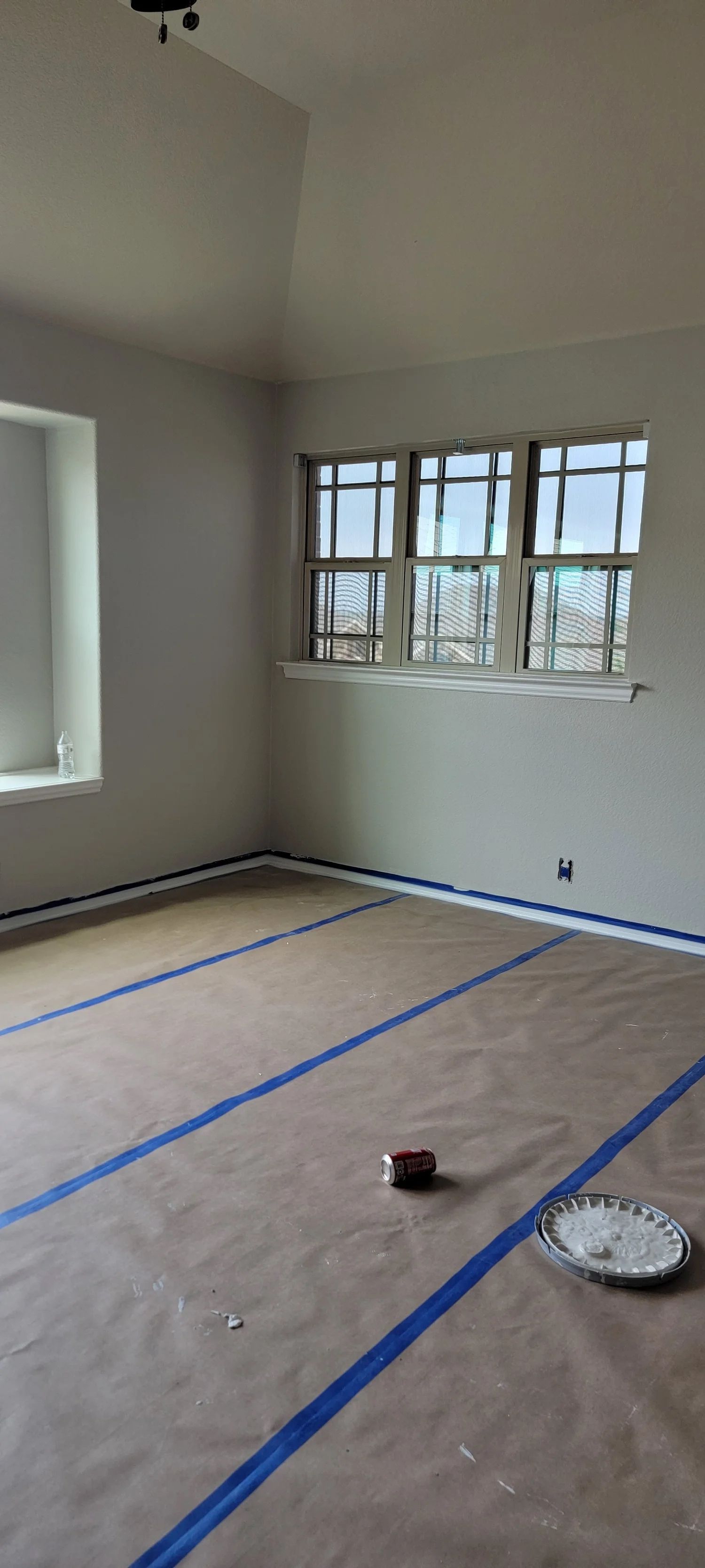 An empty room with blue painter's tape on the floor. A window on the right side and a small one on the left.