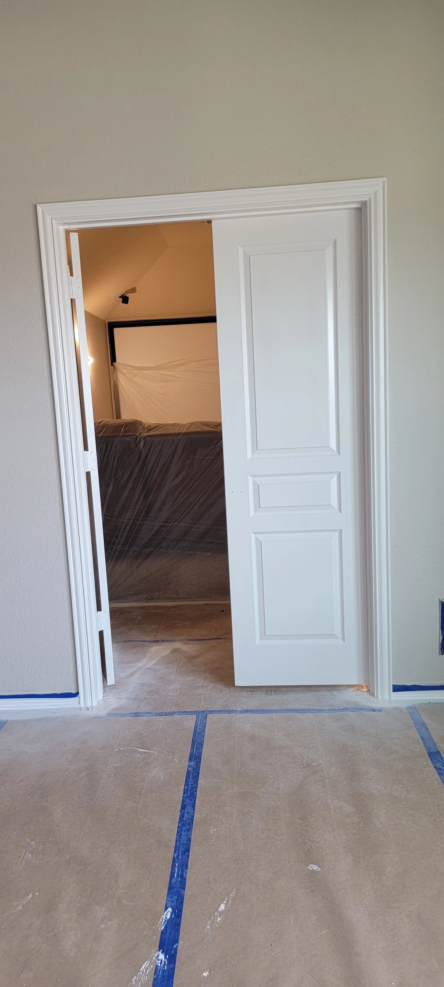 A white door partially open inside a new construction doorway.  Blue tape protects the floor.