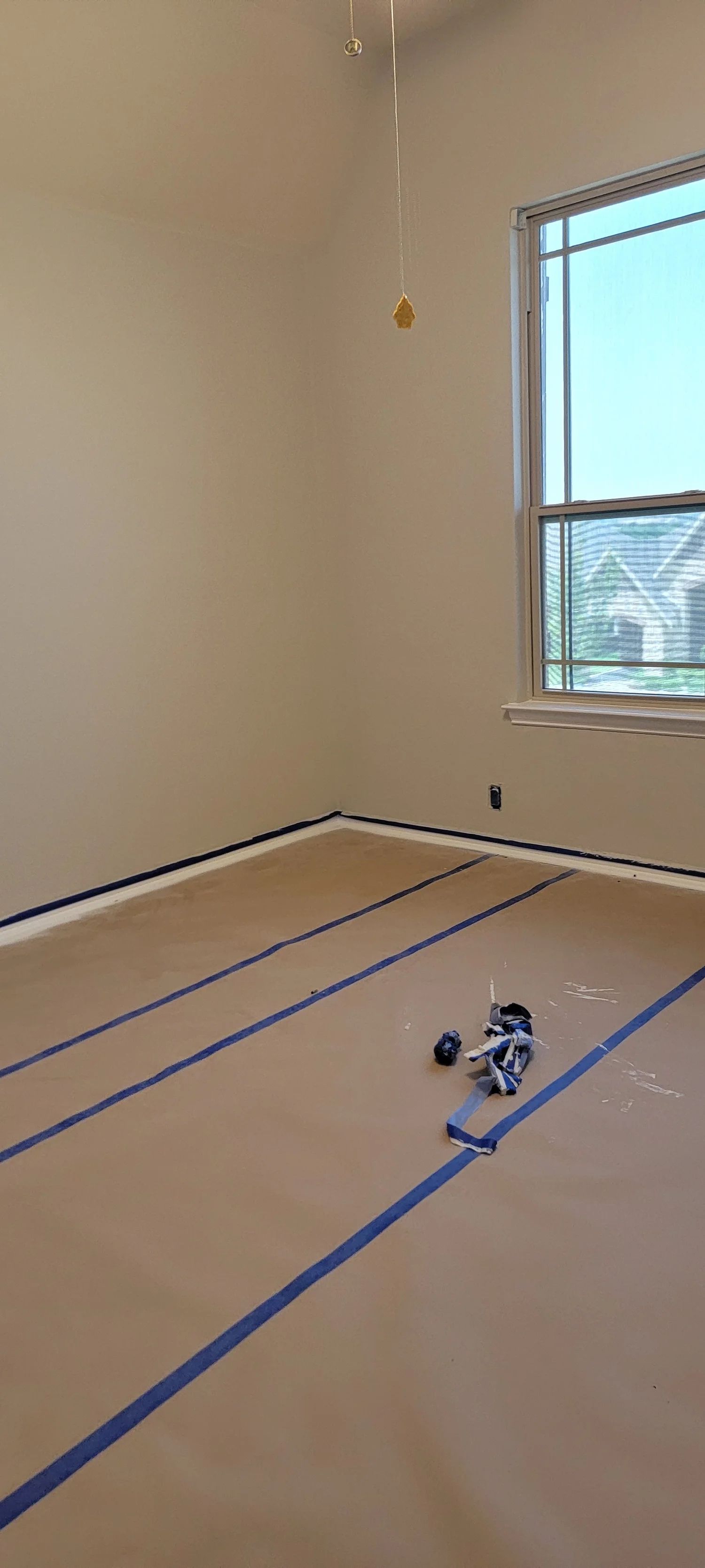 Room being painted, floor covered with paper and blue tape. Window to the right, walls are tan.
