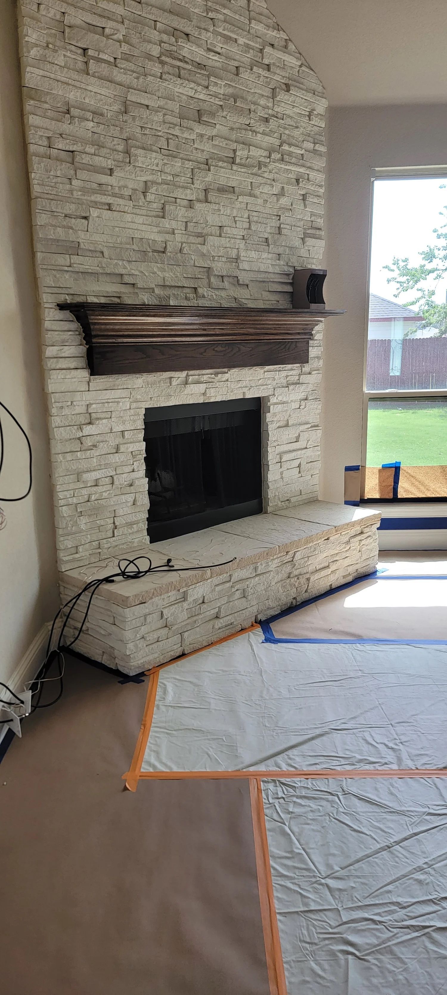 Stone brick fireplace with wooden mantel, next to a window, covered in protective materials.