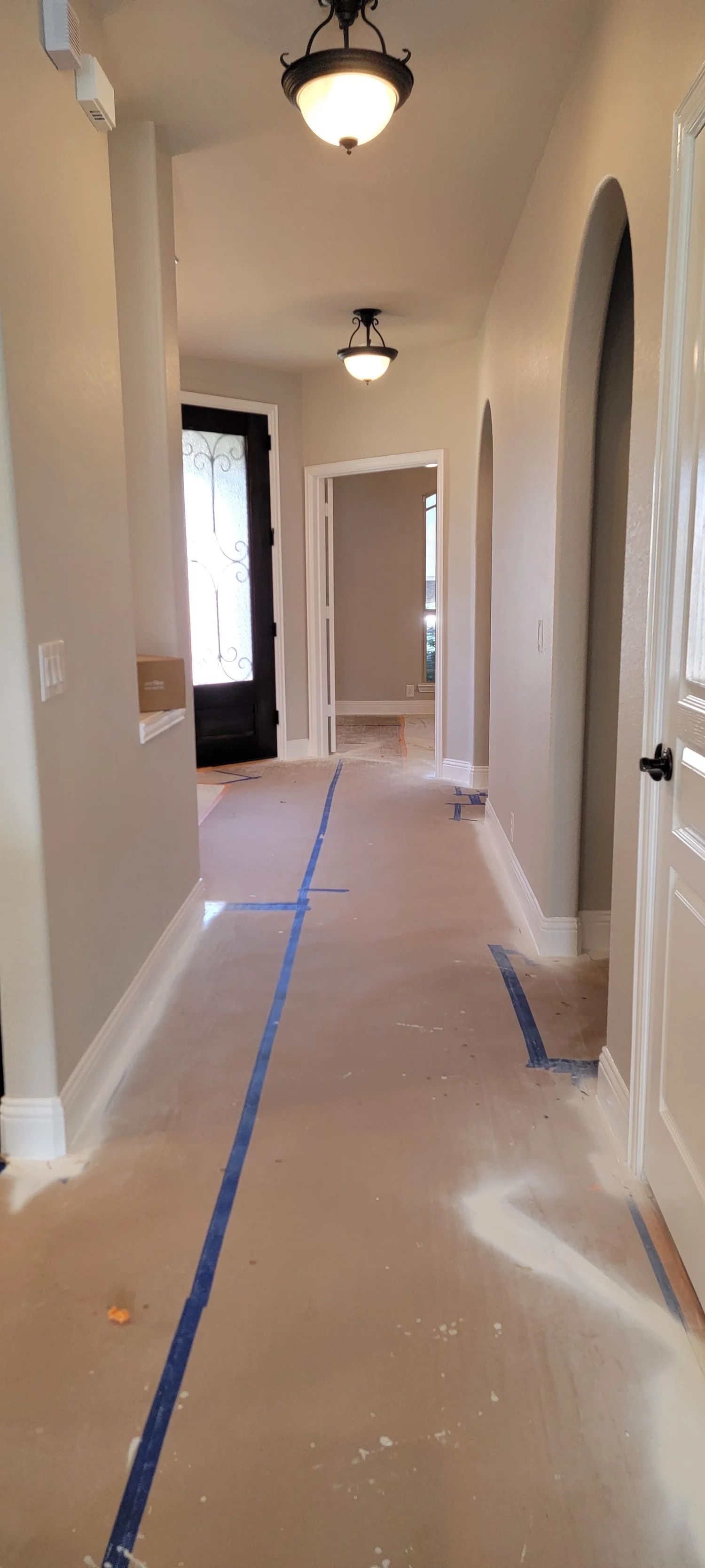 Hallway with neutral walls, trim, and brown flooring. Two pendant lights hang from the ceiling. Blue tape is on the floor.
