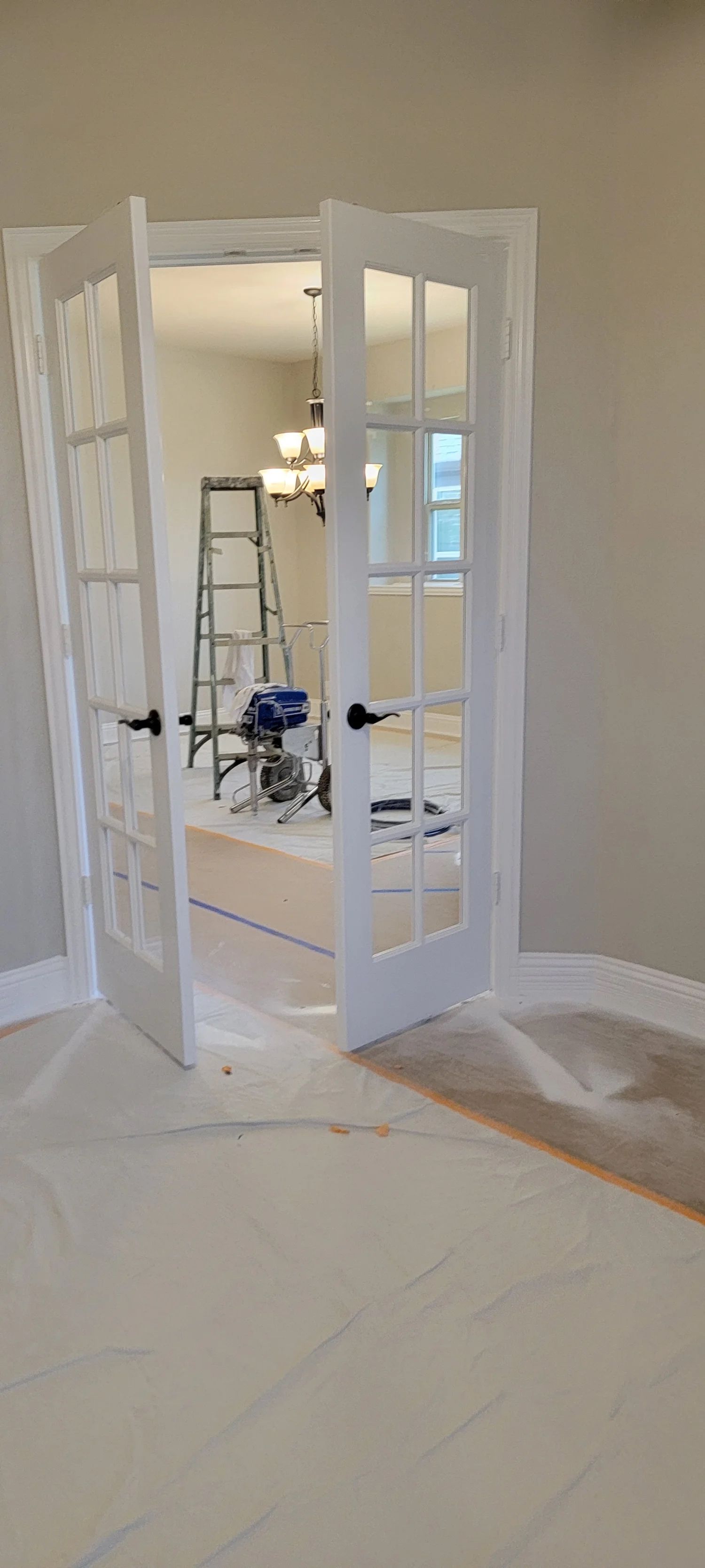 White French doors open into a room with a ladder and paint equipment. The floors are covered with protective sheeting.