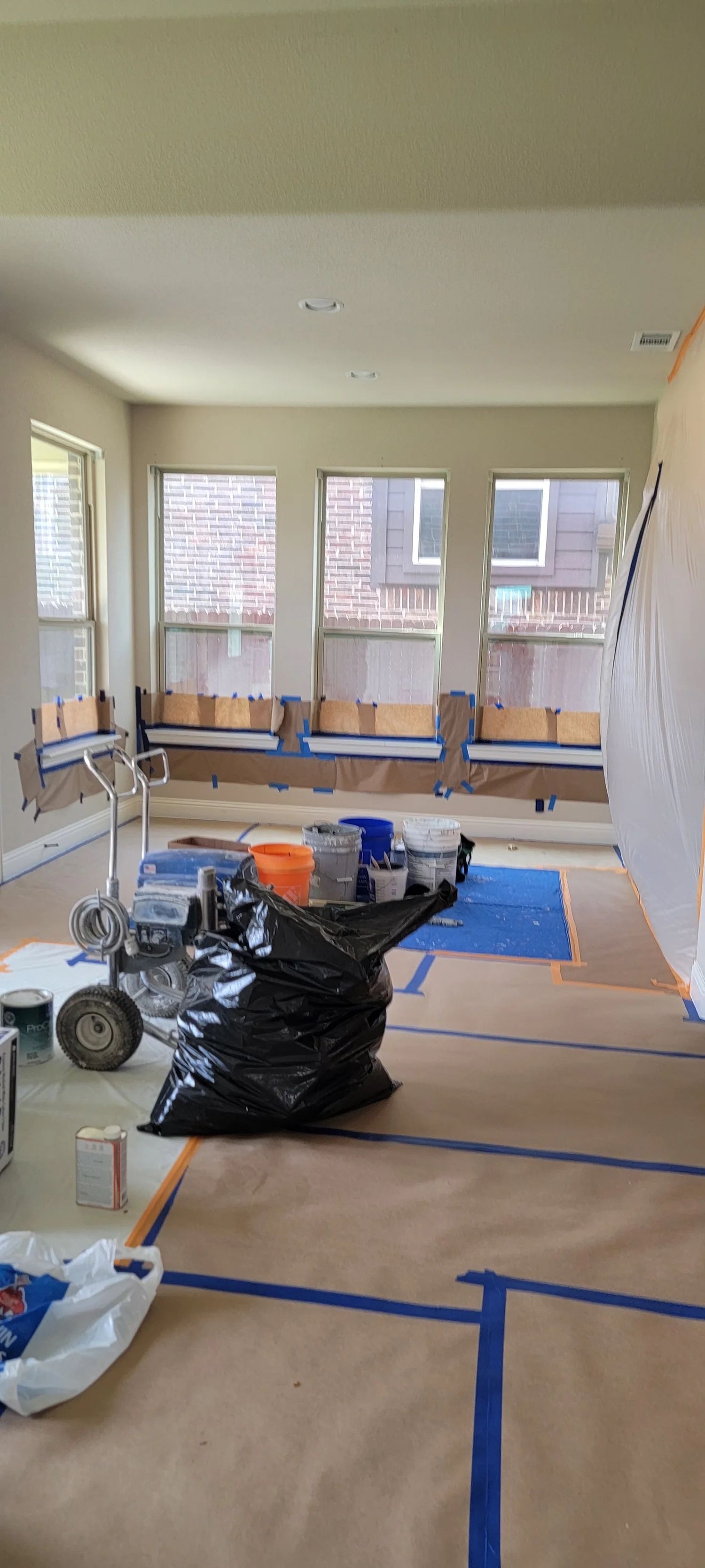 Room interior being prepped for painting: windows, painter's supplies, drop cloths, and trash bags.