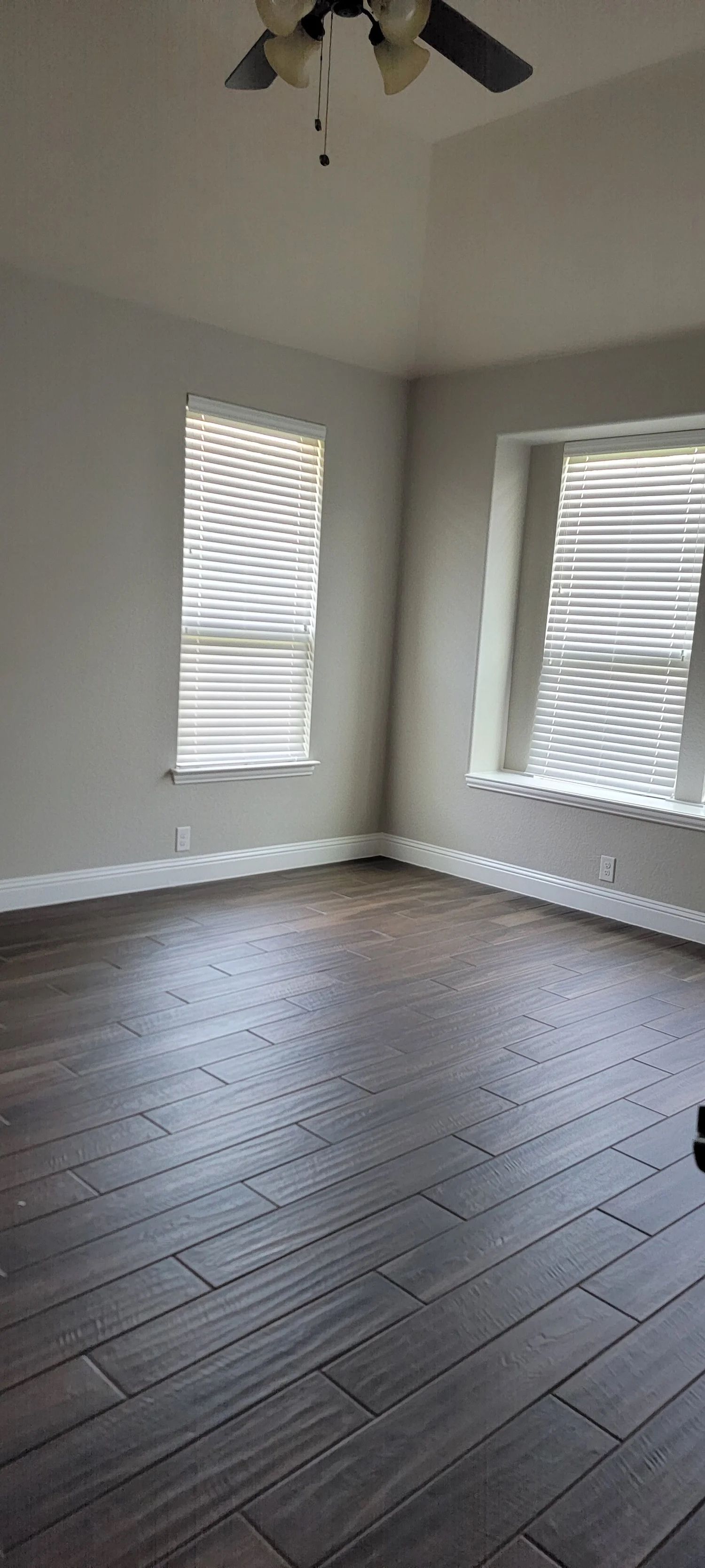 Empty room with dark wood-look tile flooring, two windows with blinds, and a ceiling fan.