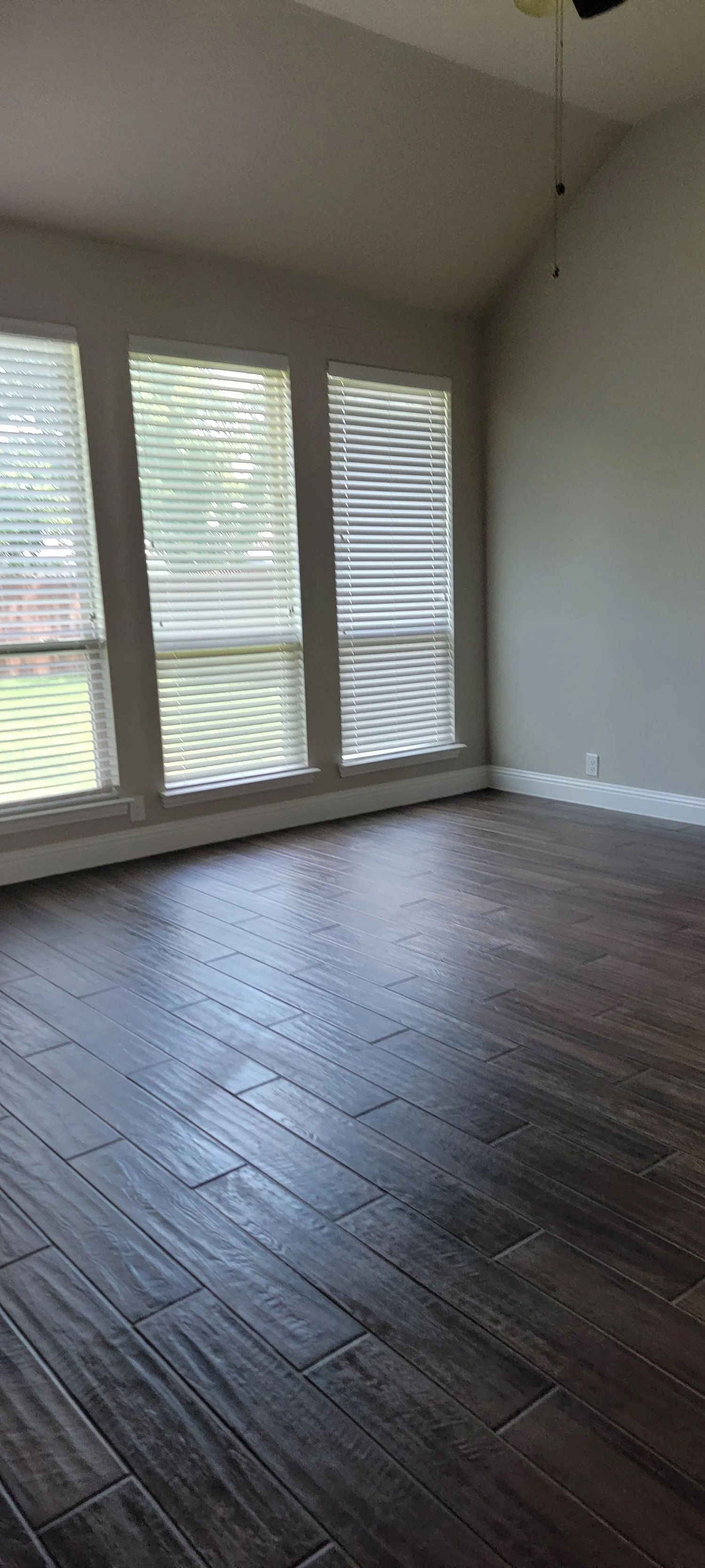 An empty room with dark wood-look tile flooring and three windows with blinds.