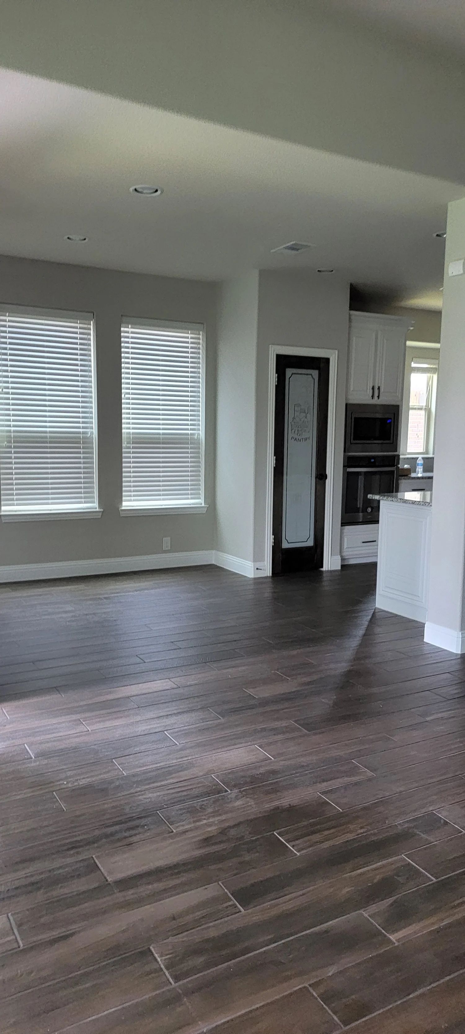 Interior view of a living space with tiled flooring and windows. A kitchen is visible in the background.