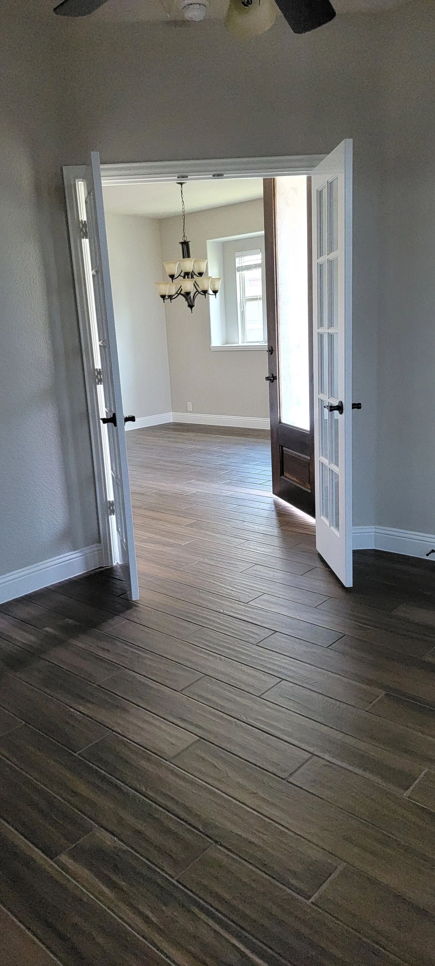 Interior view through French doors to a room with a chandelier and window. Brown floor.