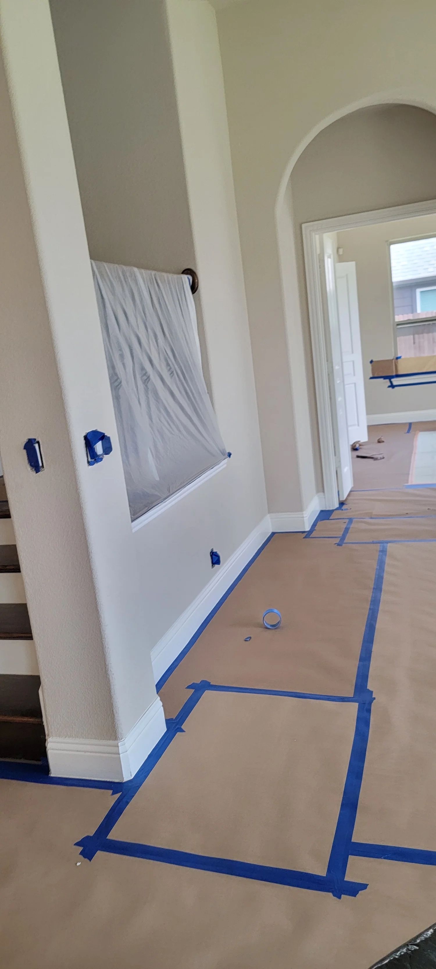 Interior room with blue tape on the floor and wall. Window covered in plastic. Arched doorway leads to another room.