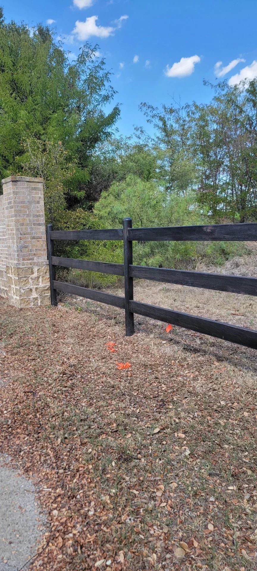 Black wooden fence next to a stone pillar, with trees and a blue sky in the background.
