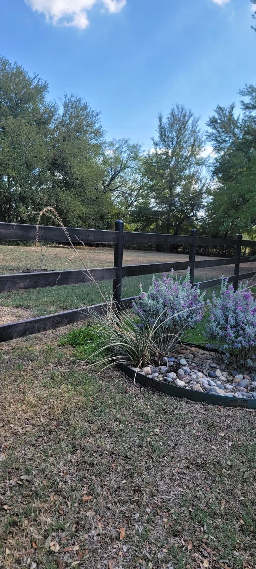 A split-rail fence separates a lawn with landscaping from trees under a blue sky.