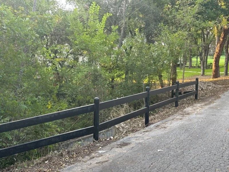 Black two-rail fence along a paved road, green foliage in the background.