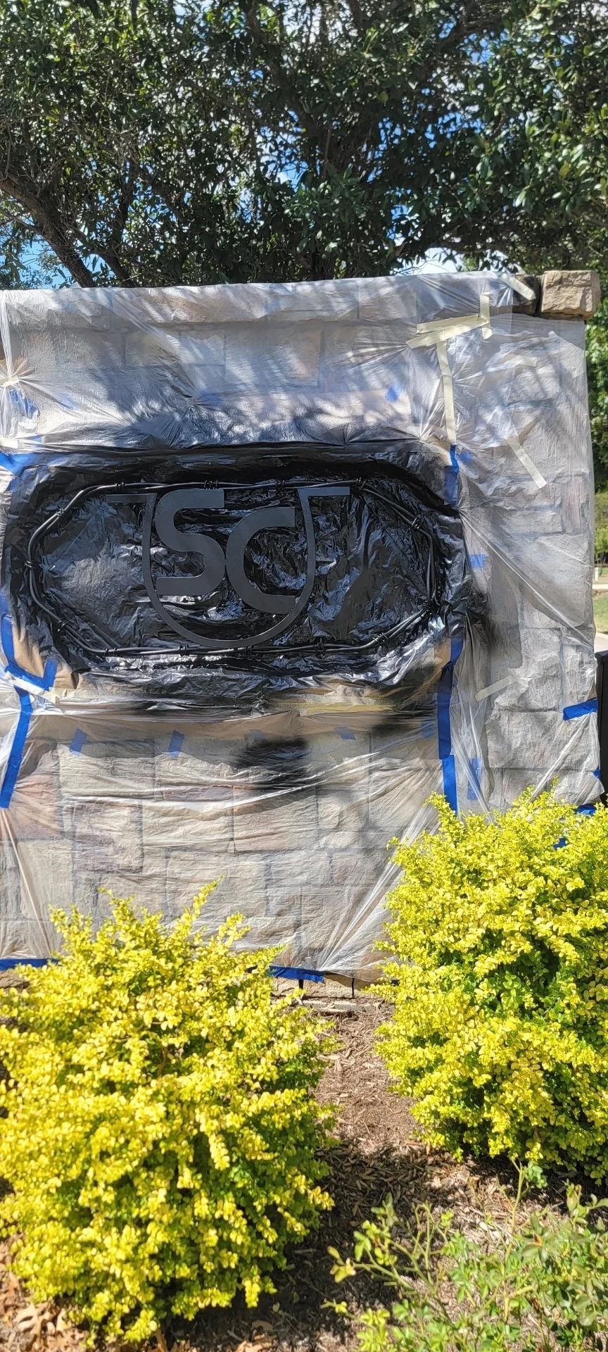 Black painted sign on brick wall, covered in plastic and tape, flanked by yellow bushes.