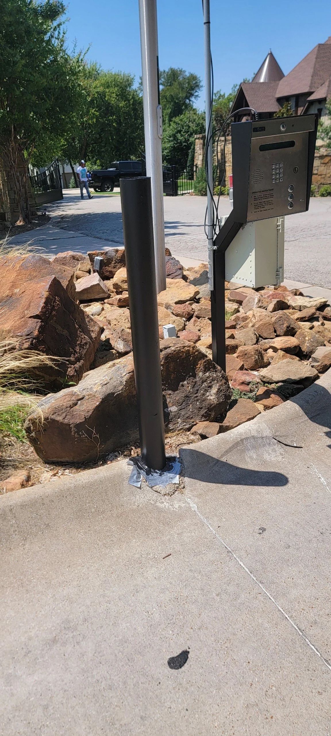 Mailbox on a post, set among rocks next to a sidewalk and a tree line under a clear sky.
