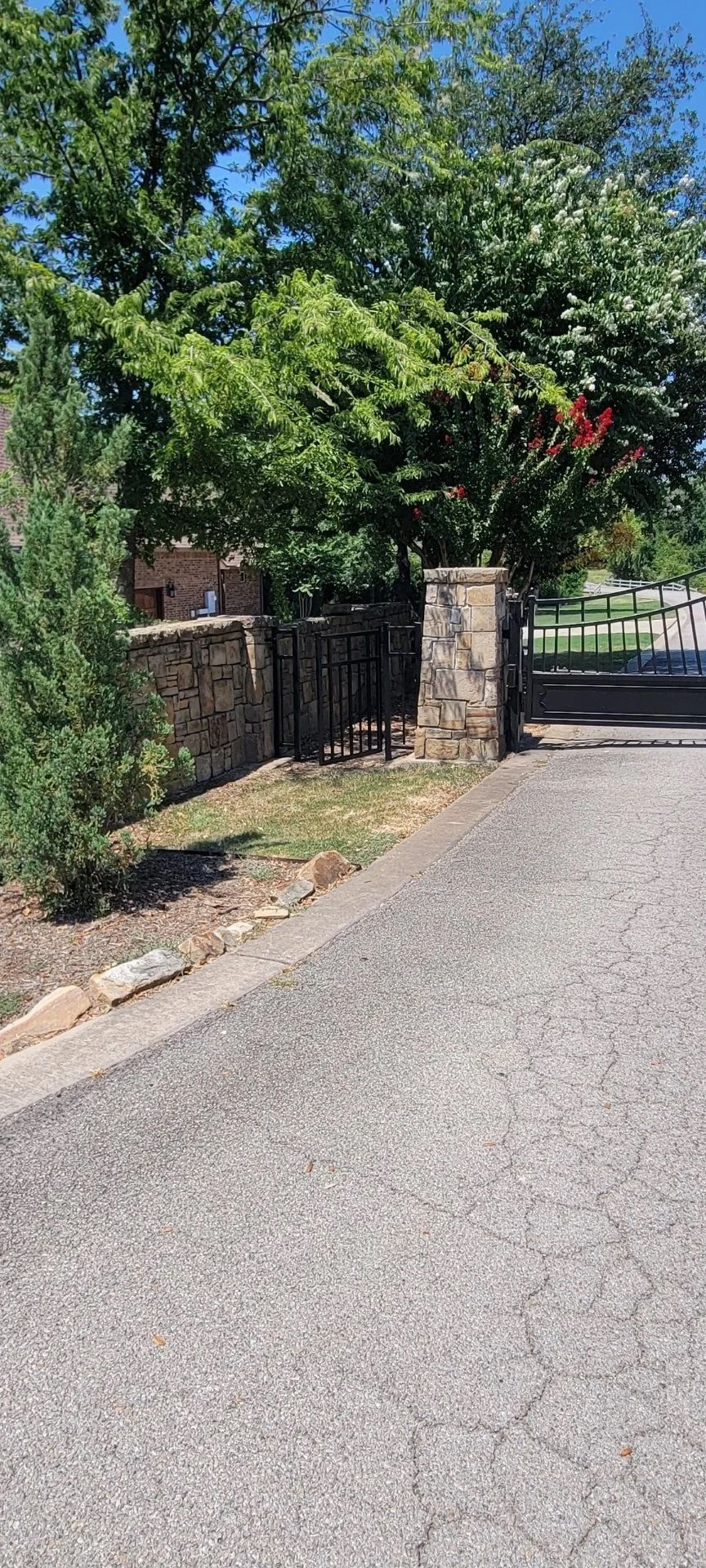 Stone gate entrance with iron gates, surrounded by trees and a paved road.