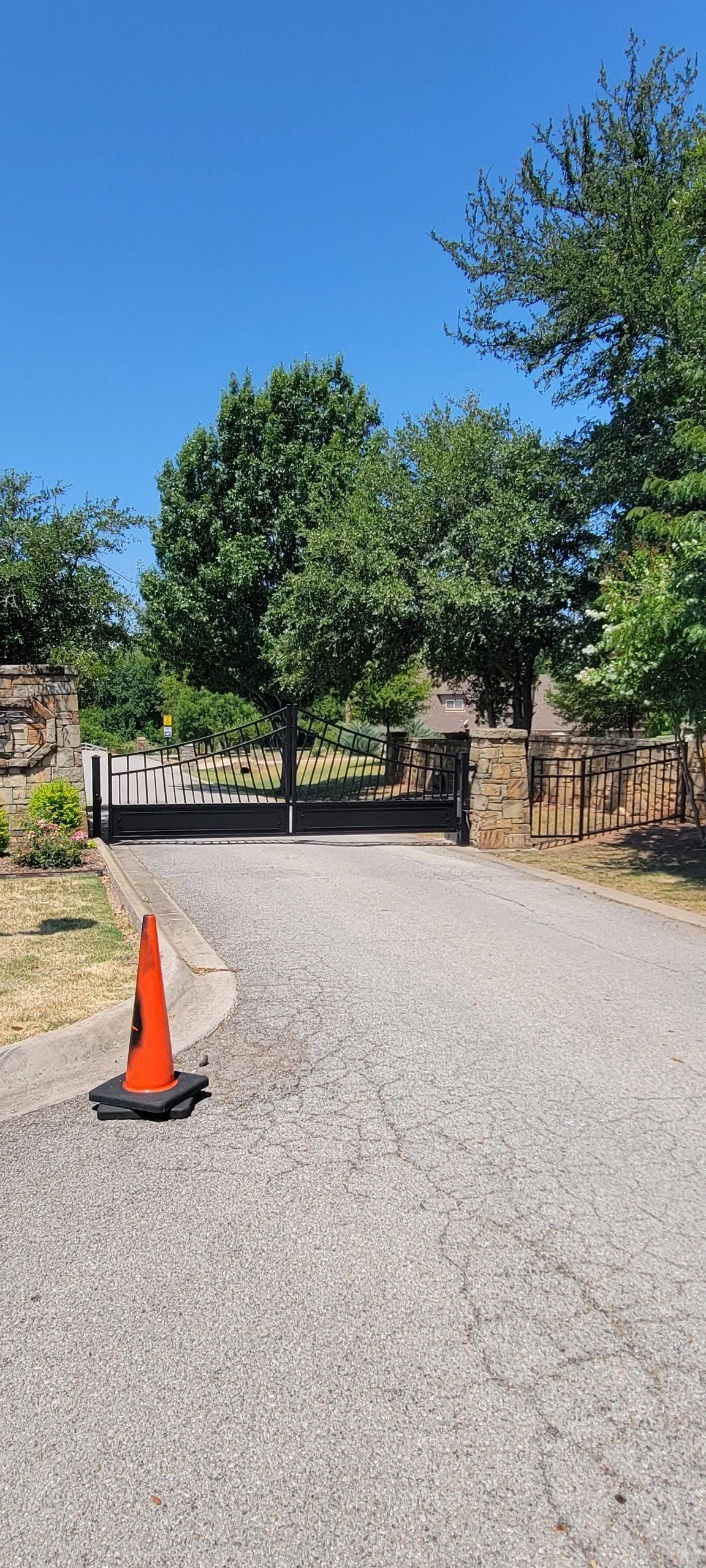 Orange traffic cone on a gravel road, leading to a gate framed by trees and blue sky.