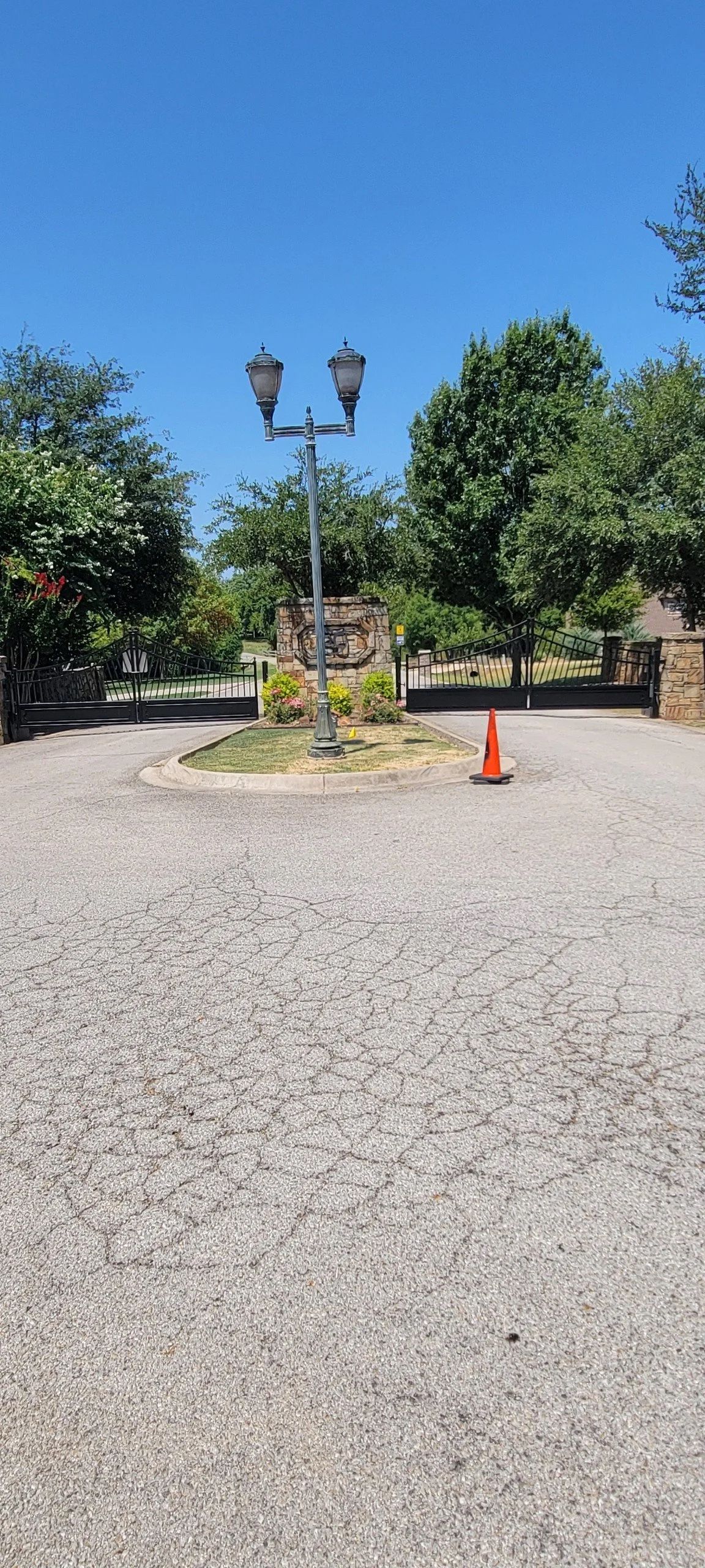 Gravel road leads to a lamp post and small garden, an orange cone to the right. Green trees and blue sky background.