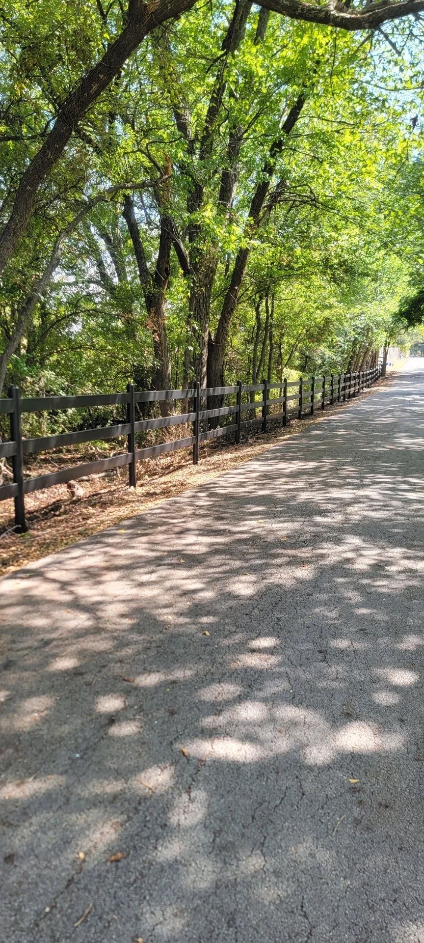 Gravel road lined by a wooden fence and trees casting shadows. Sunlight filters through the leaves.