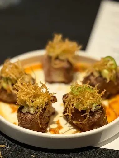 Five seared steak bites topped with fried onions, sauce, and a slice of jalapeño, served on a white plate.