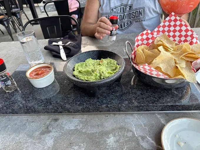 Guacamole, chips, and salsa served on a stone platter at an outdoor restaurant. A person adds pepper.