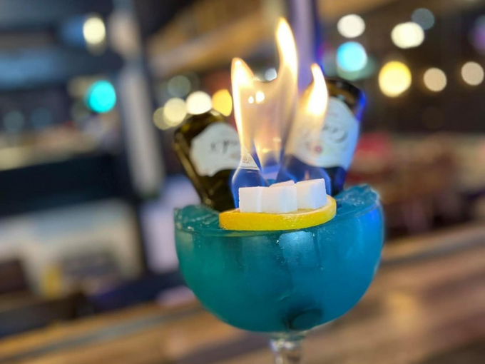 Blue flaming cocktail garnished with lemon and sugar cubes, two bottles in the background.