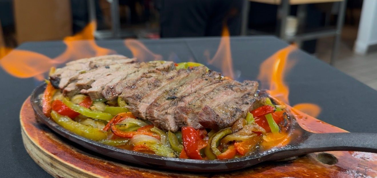 Sizzling steak fajitas on a hot plate, with flames rising. Red and green peppers and onions visible.
