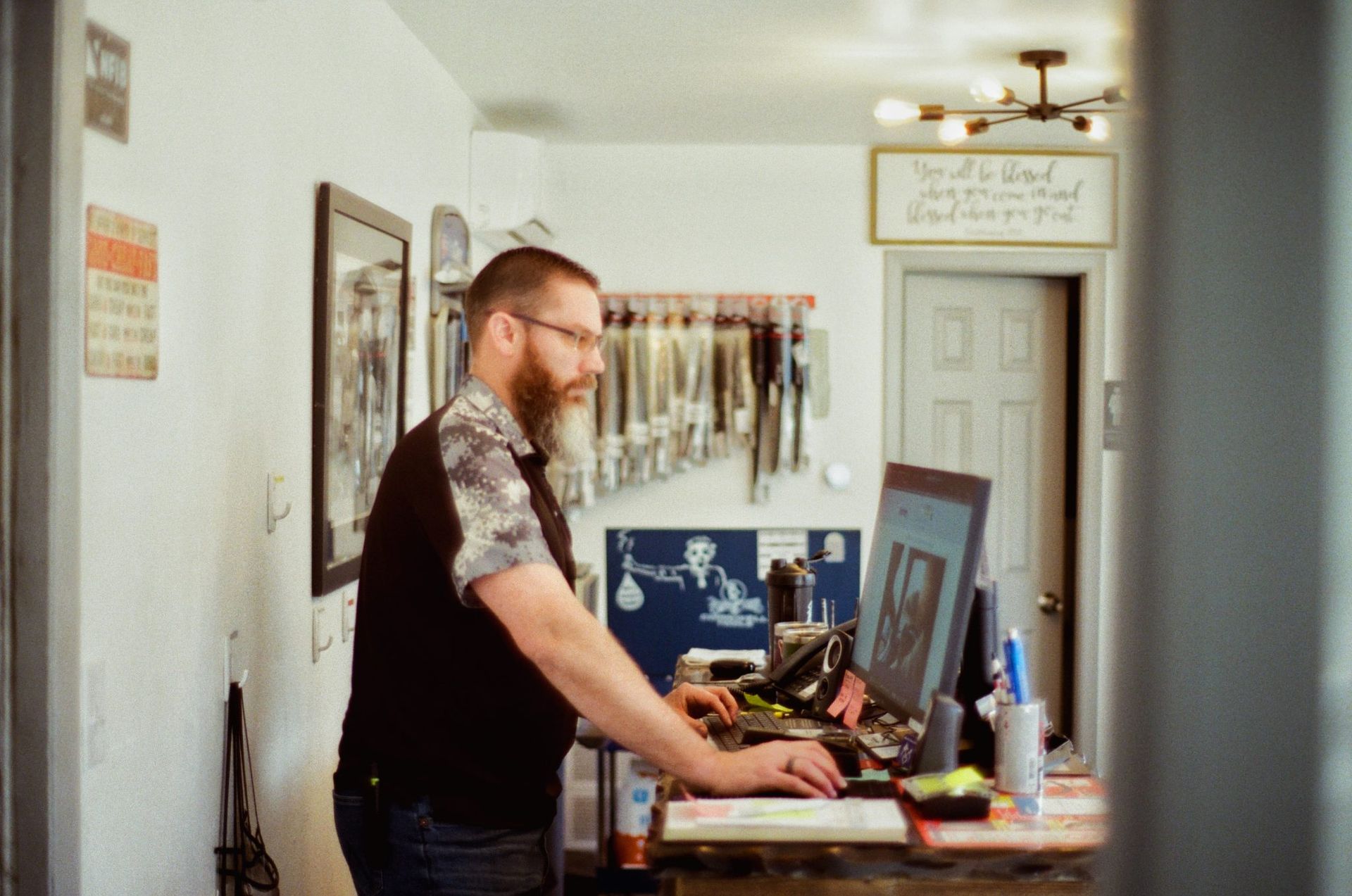 Man standing at a desk in a bright hallway, working on a computer surrounded by tools and framed art. | Buck & Doe Auto