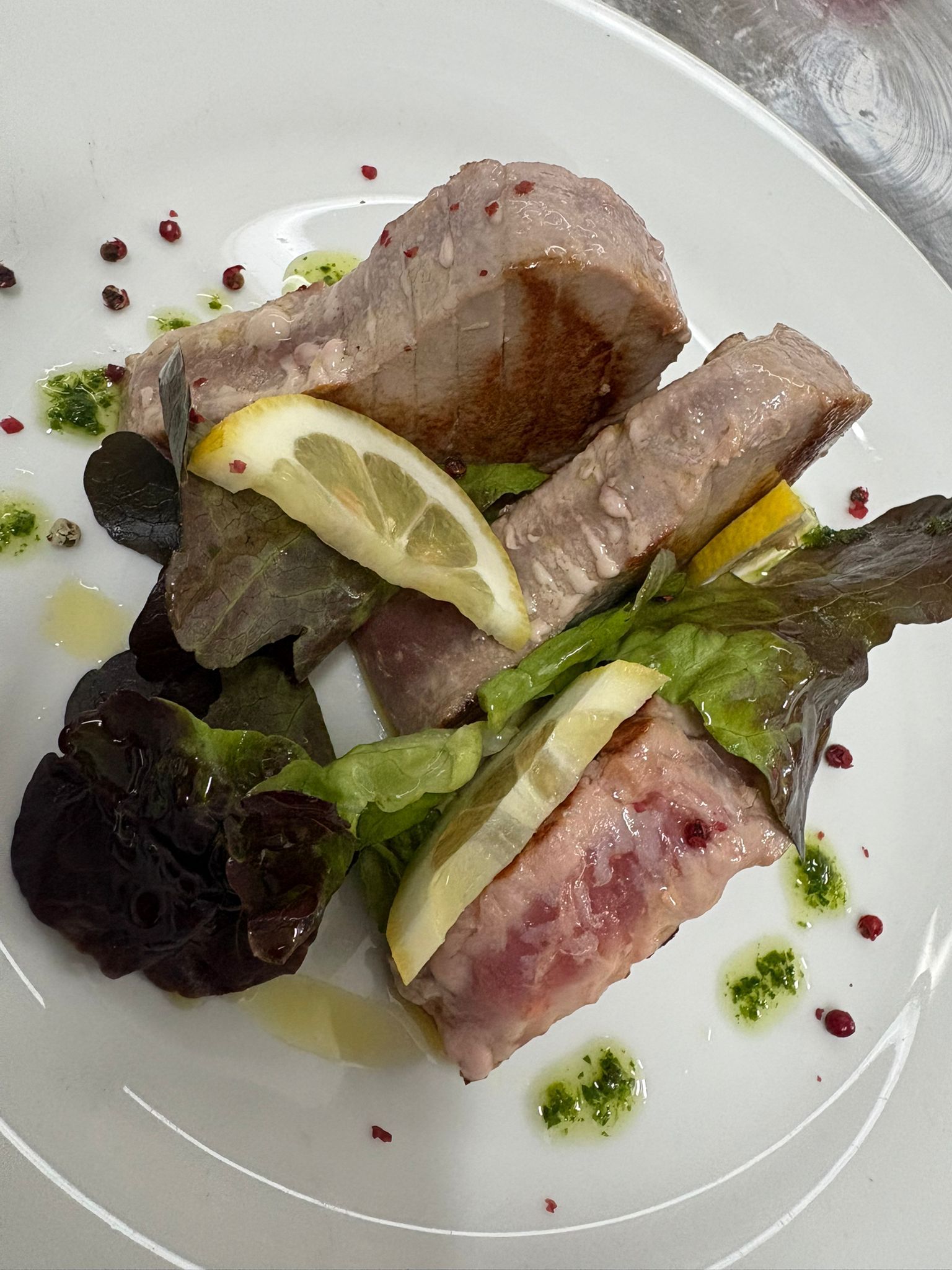 Piatto di pesce gourmet impiattato con spicchi di limone, verdure e guarnizione di erbe aromatiche su un piatto bianco.