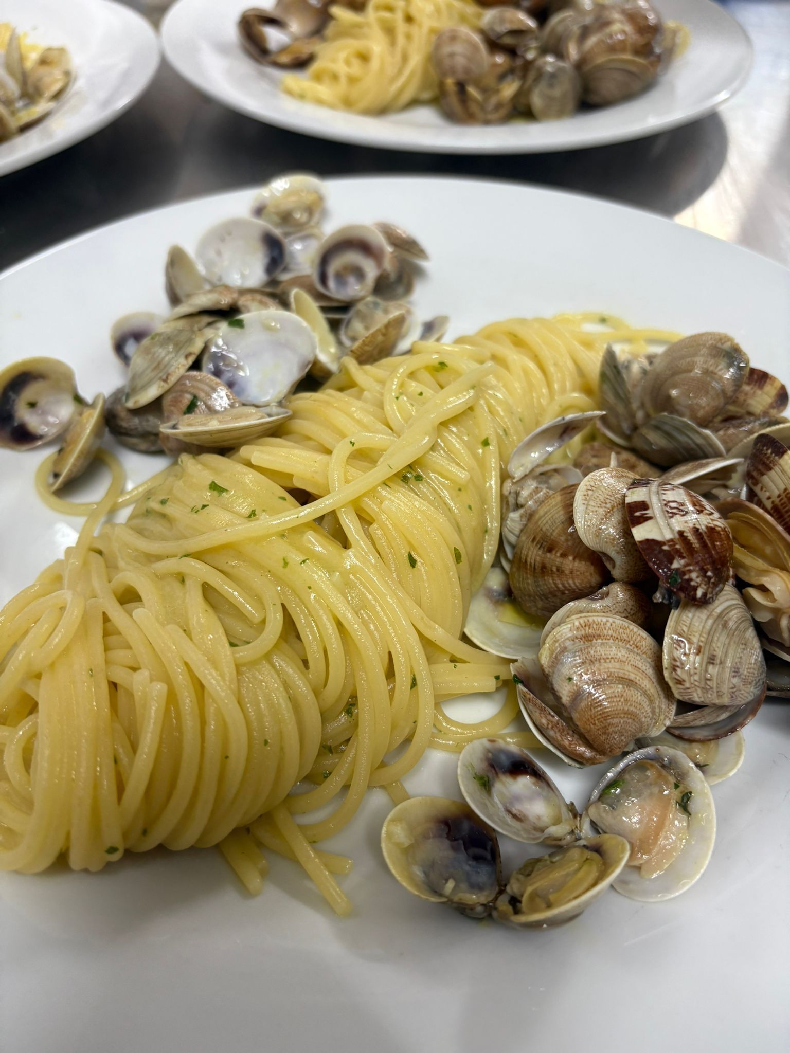 Un piatto di spaghetti al sugo di vongole su un tavolo bianco, con altri due piatti sullo sfondo.
