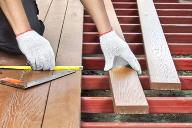 A person wearing white gloves measures and installs brown wood-like decking planks onto a red metal frame.