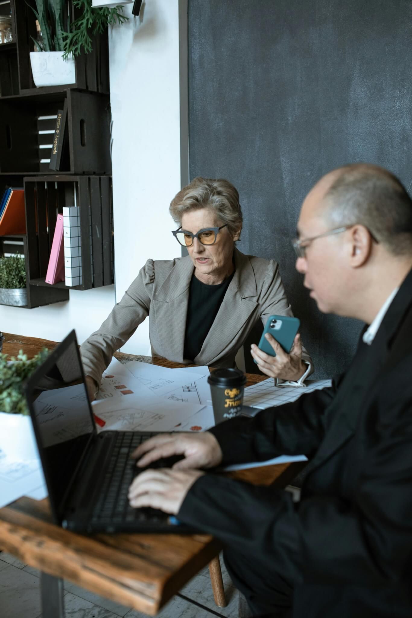 Mulher e homem em uma mesa, olhando para um laptop e papéis, discutindo trabalho.
