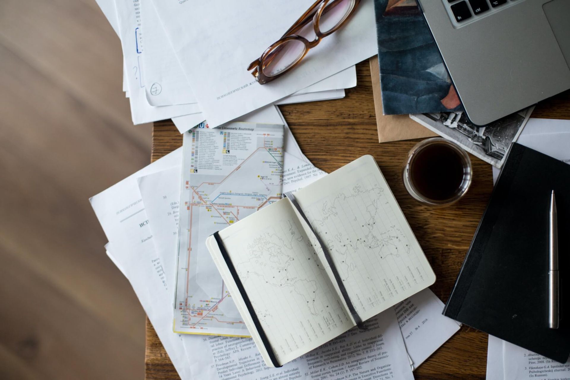 Mesa cheia de papéis, óculos, laptop, caderno, mapa e café.