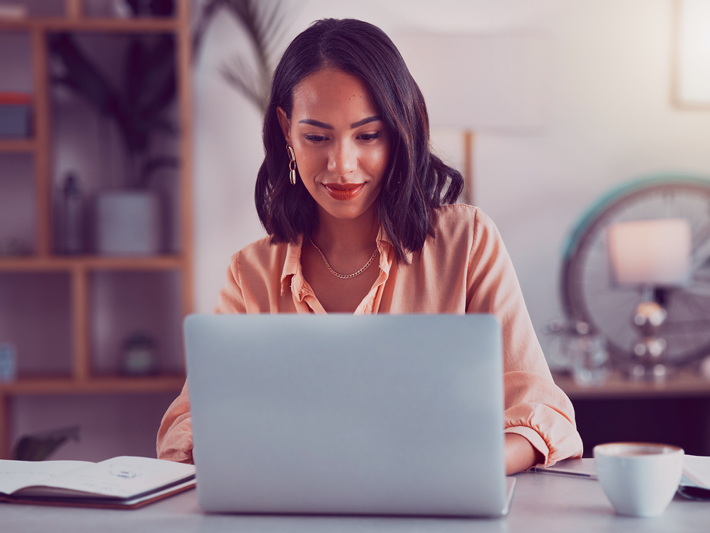Mulher trabalhando em um laptop em uma mesa com café, livros e plantas.