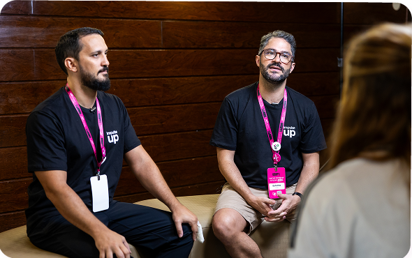 Dois homens de camisa preta e crachá rosa estão sentados em uma mesa, conversando com alguém em primeiro plano.
