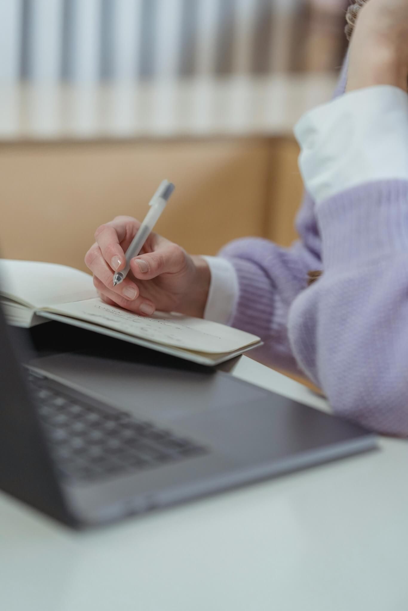 Pessoa escrevendo em um caderno, suéter lilás, perto de um laptop.