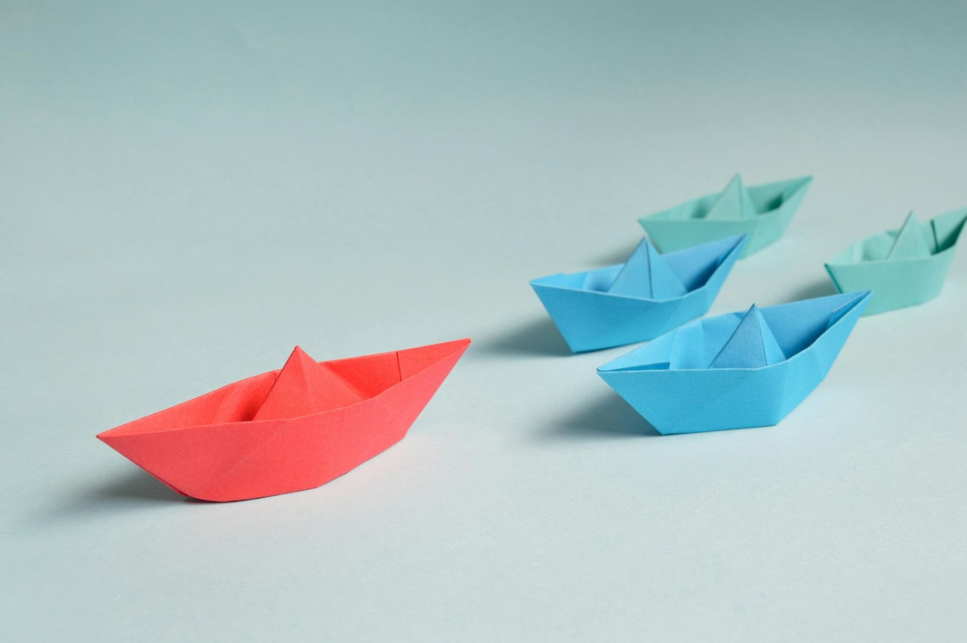 Barco de papel vermelho liderando uma fileira de barcos de papel azul e verde-azulado em uma...