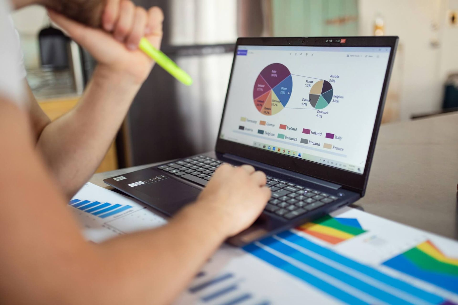 Pessoa trabalhando em um laptop, revisando gráficos de pizza e barras, segurando um marca-texto 