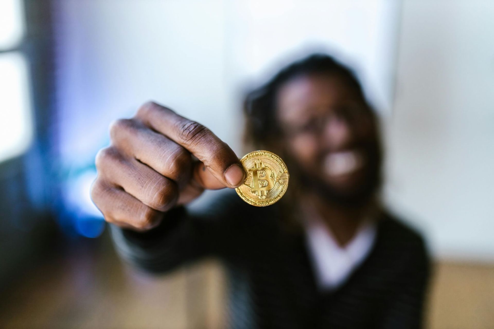 Homem negro sorrindo segurando uma moeda dourada de Bitcoin. Fundo desfocado.