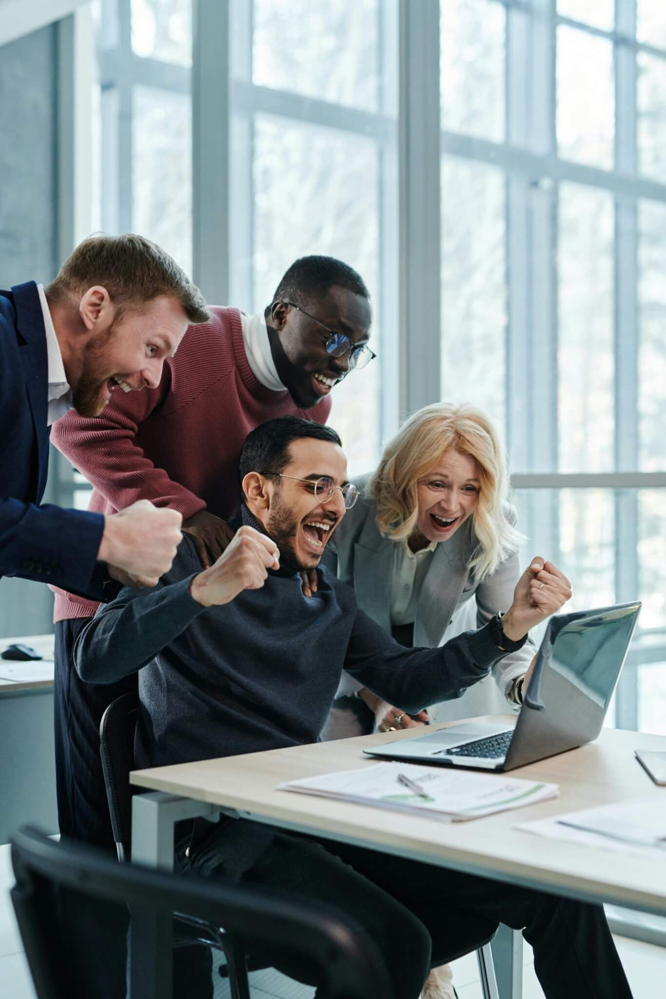 A equipe comemora em volta de um laptop, sentados à mesa. Eles estão diversos, animados e felizes, em um escritório.