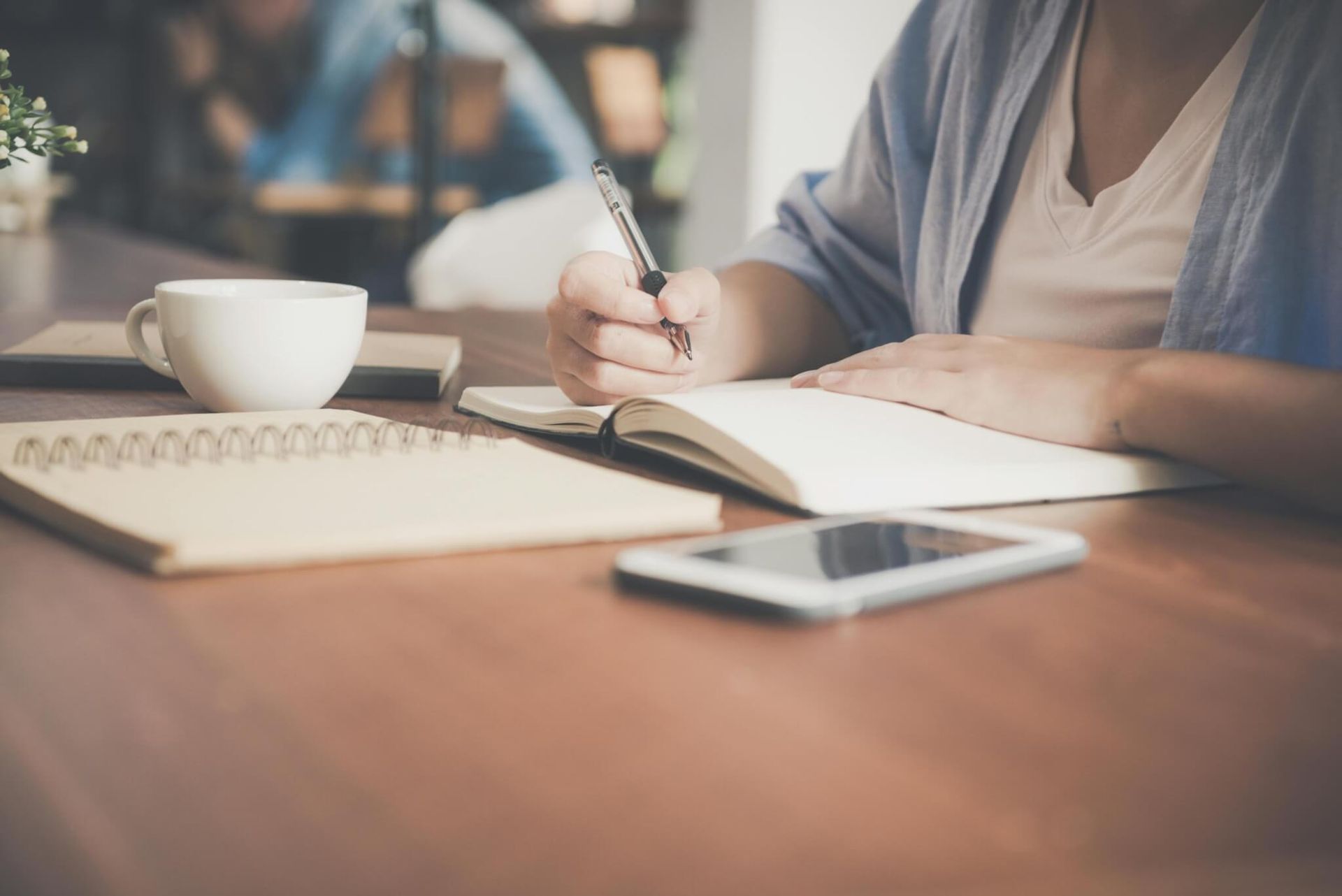 Mulher escrevendo em um caderno em uma mesa com uma xícara de café, telefone e outro caderno.